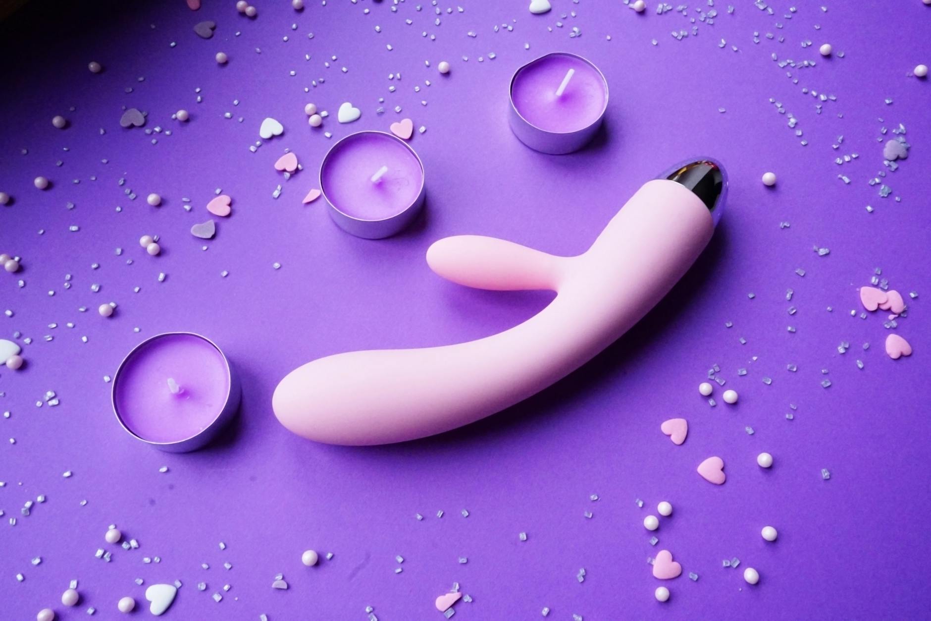 Pink vibrator on a purple background with heart confetti and candles symbolizing intimate self-care after heartbreak.
